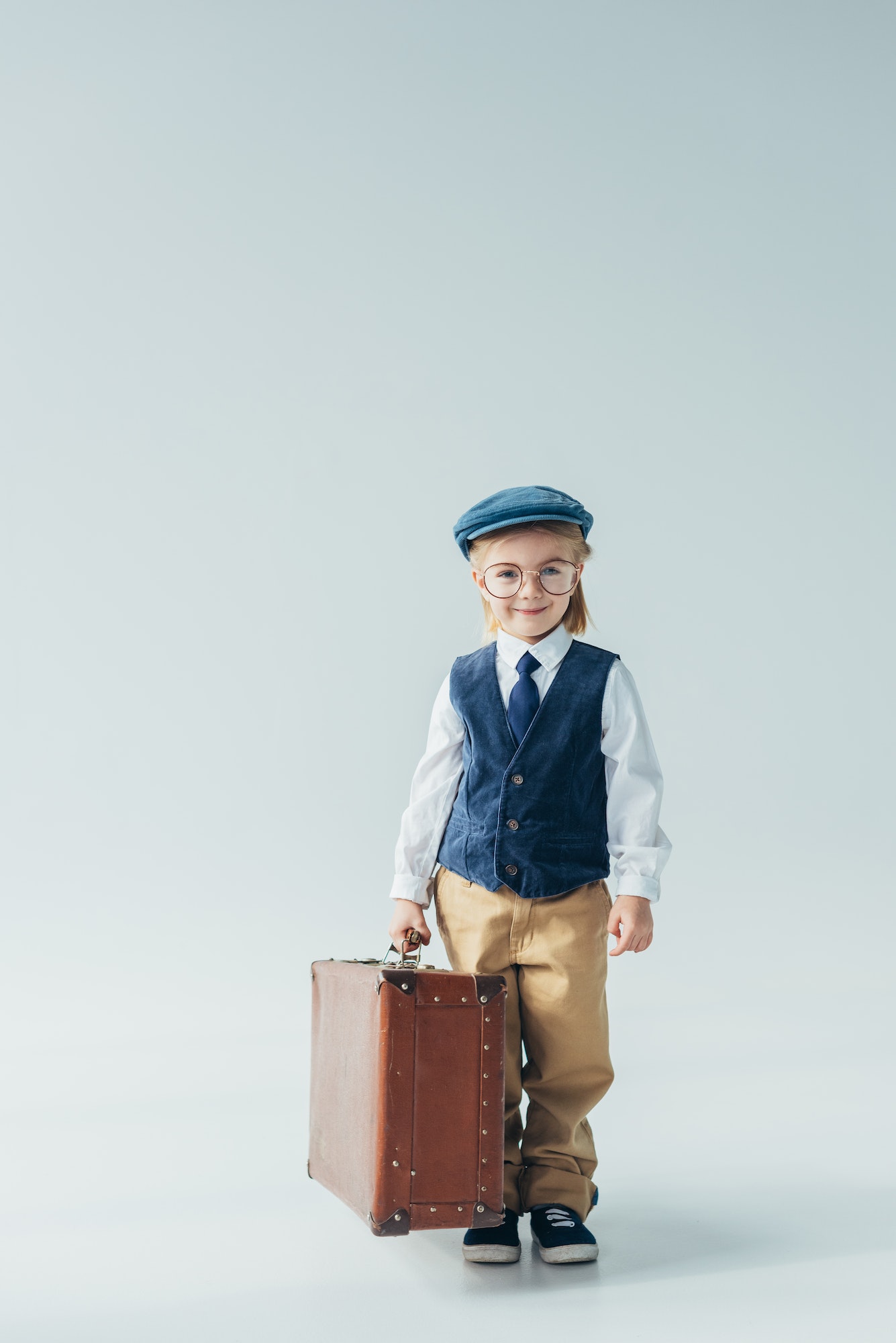 happy kid in retro vest and cap holding suitcase and looking at camera